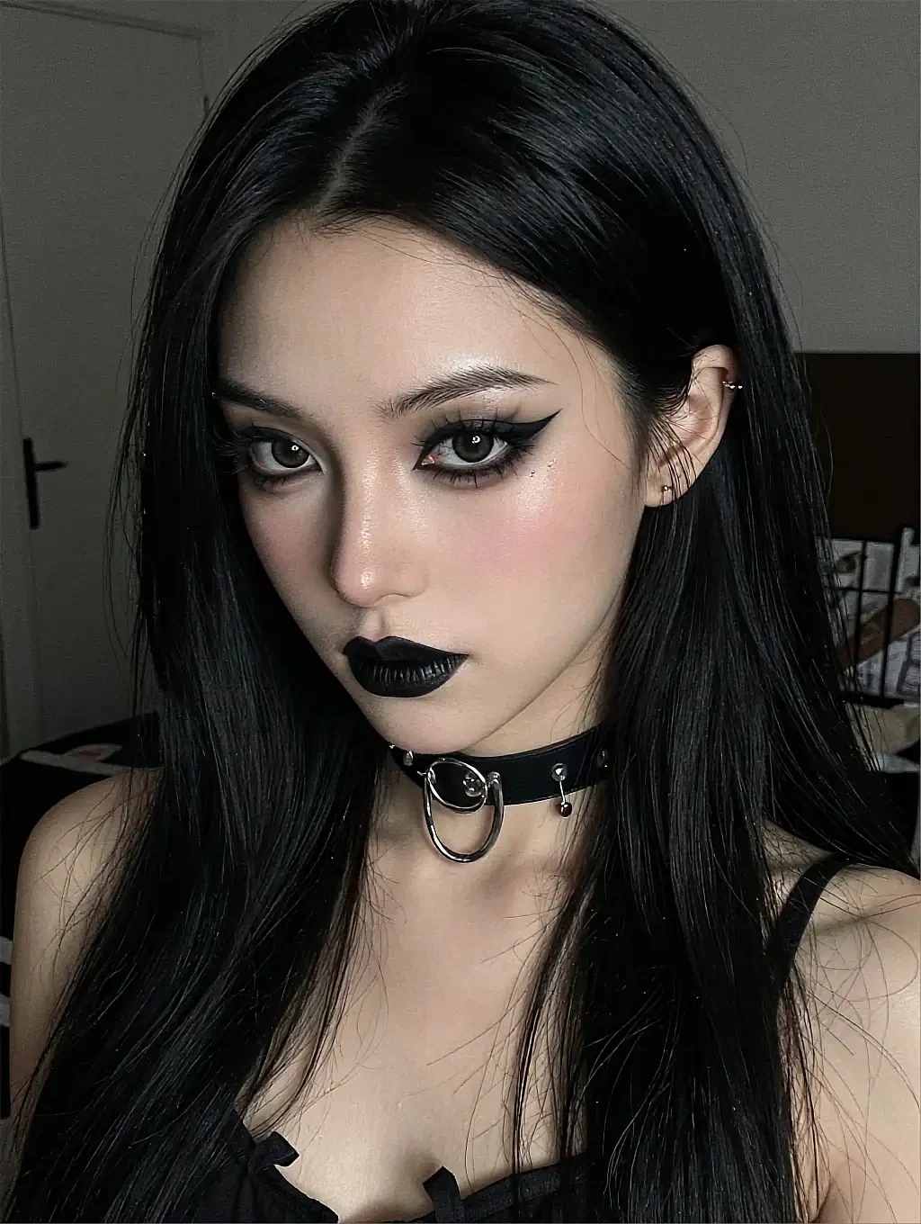 Fictional AI character with black hair, black lipstick and a studded choker in a strappy black top in a dim room